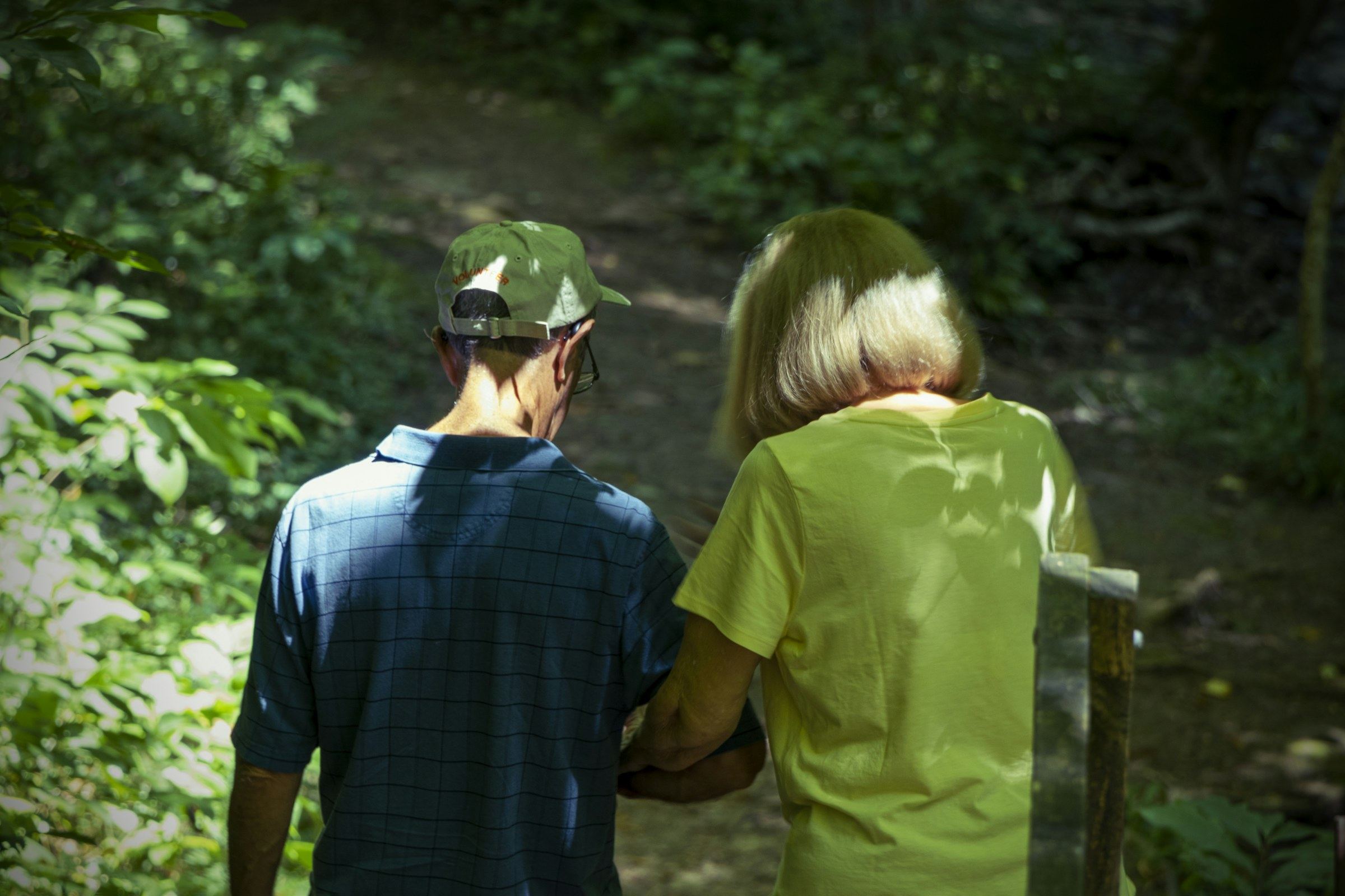 An adult walking arm in arm with an elderly parent through a wooded path